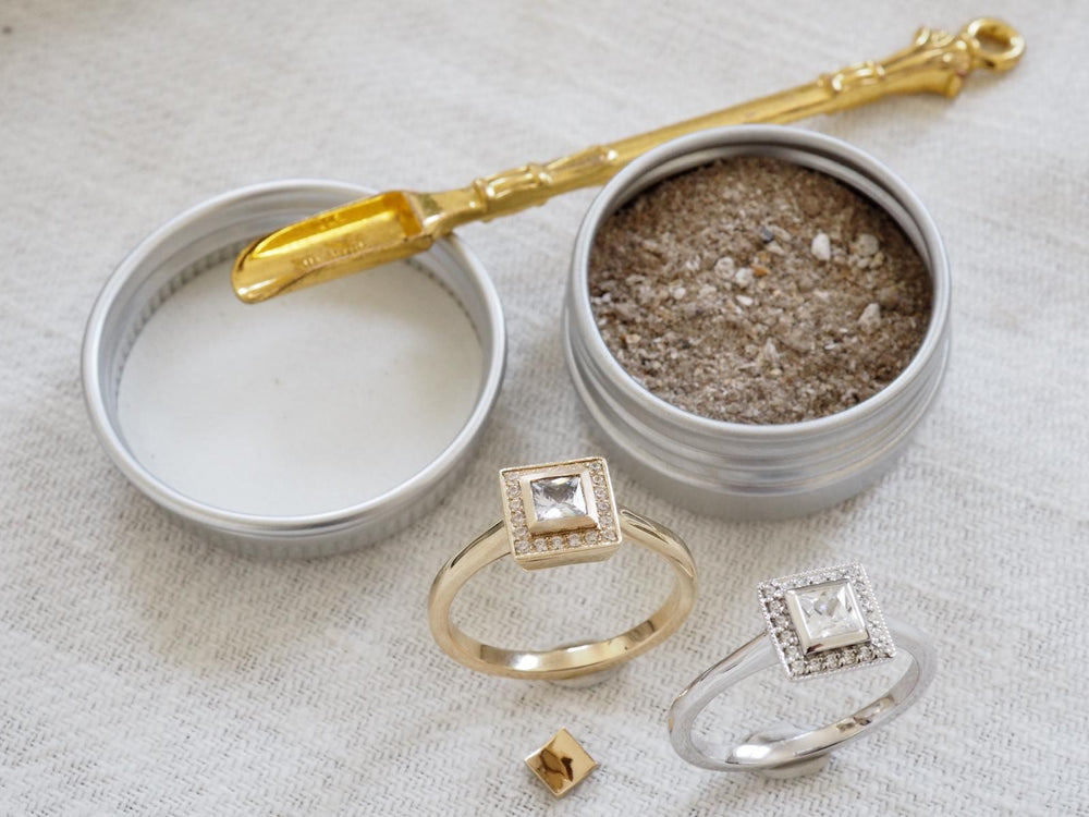 Two round metal tins containing cremation ashes, with a gold-colored spatula resting across them, alongside two sample rings on a textured white fabric background.